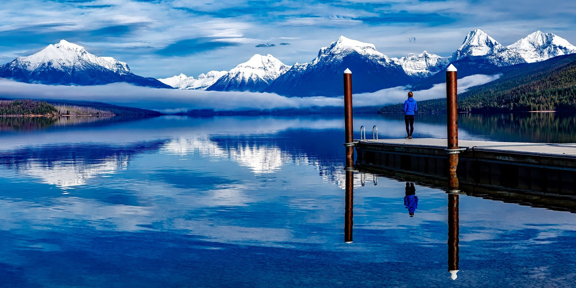 Person standing on a dock with snow-capped mountains and a clear blue sky.