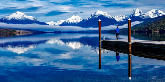 Person standing on a dock with snow-capped mountains and a clear blue sky.
